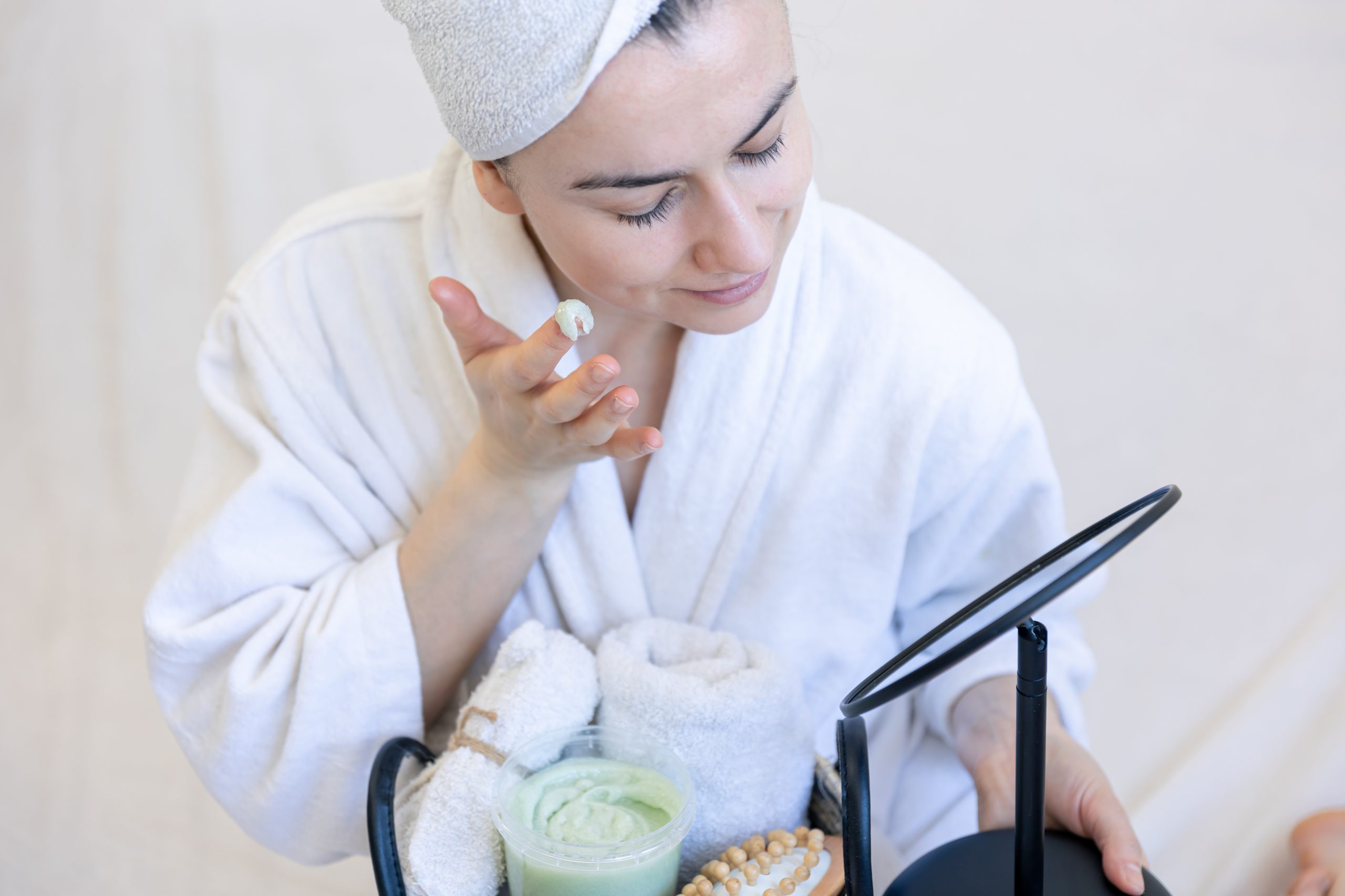 Young woman applying cream on her face, looking in the mirror, facial skin care concept, moisturizing.